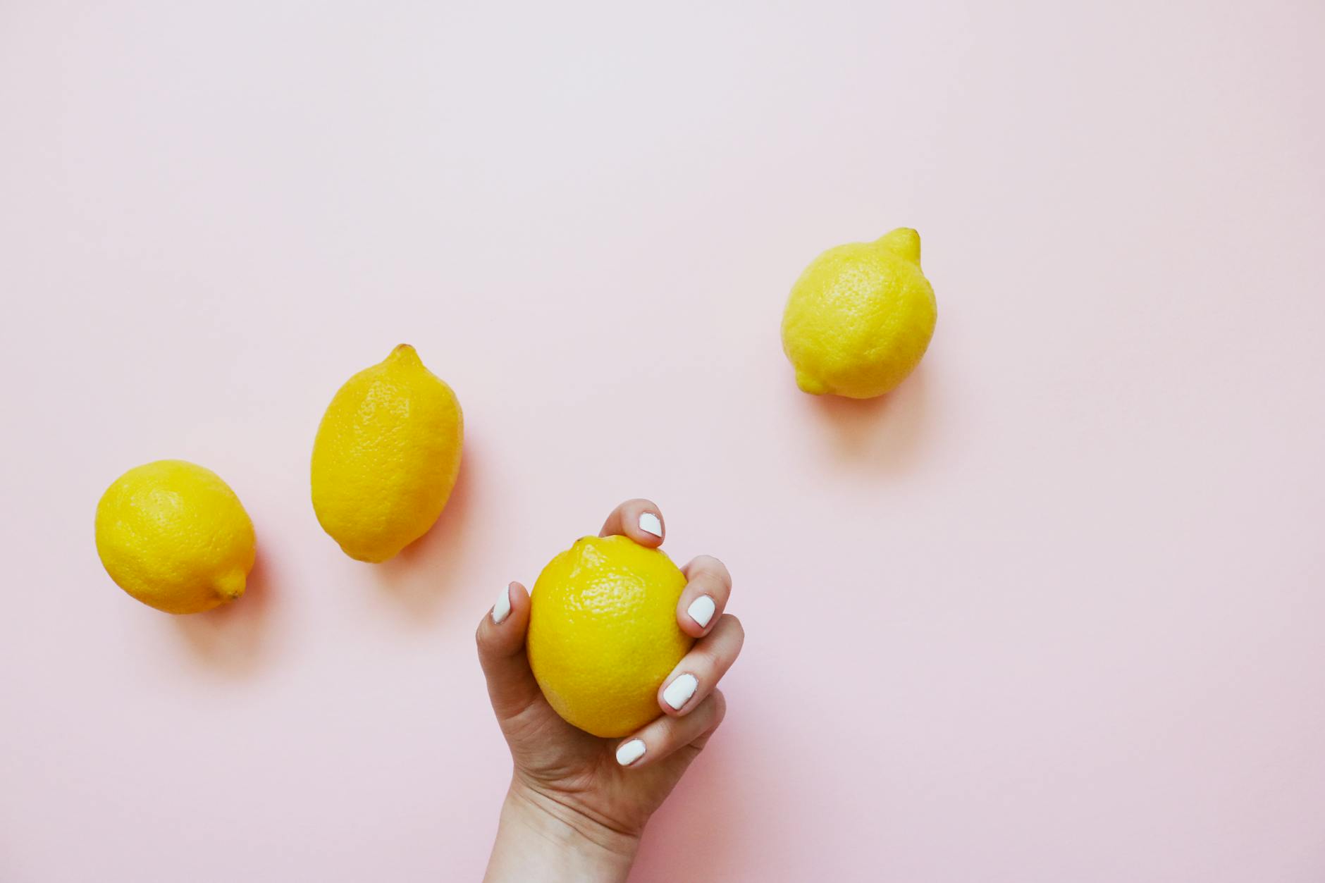 A hand with white nails holding a lemon on a soft pink background, surrounded by additional lemons