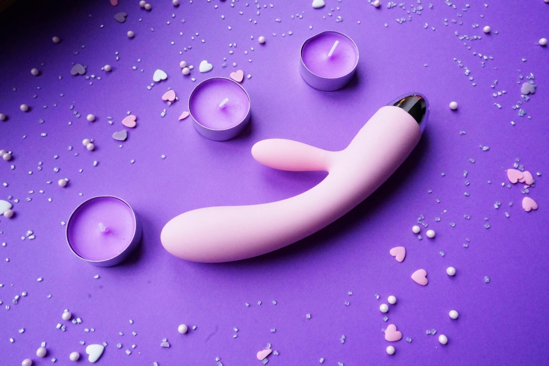 A pink vibrator on a purple background with heart confetti and candles, creating a relaxed solo pleasure atmosphere.